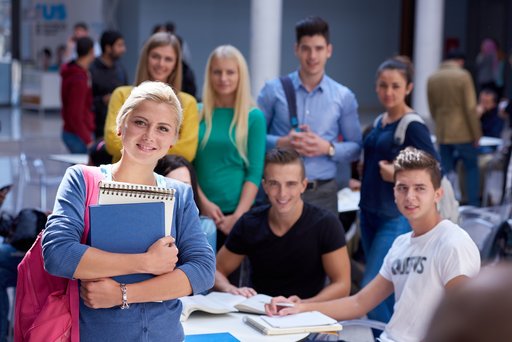 Eine junge Frau mit Büchern im Arm lächelt in die Kamera. Im Hintergrund sind weitere junge Erwachsene, die zusammen an einem Tisch sitzen und lernen.