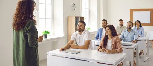Eine Szene aus der Erwachsenen-Bildung. Eine Lehrerin steht vorn. An Tischen sitzen sechs Erwachsene und hören zu.
