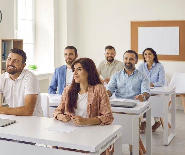 Eine Szene aus der Erwachsenen-Bildung. Eine Lehrerin steht vorn. An Tischen sitzen sechs Erwachsene und hören zu.