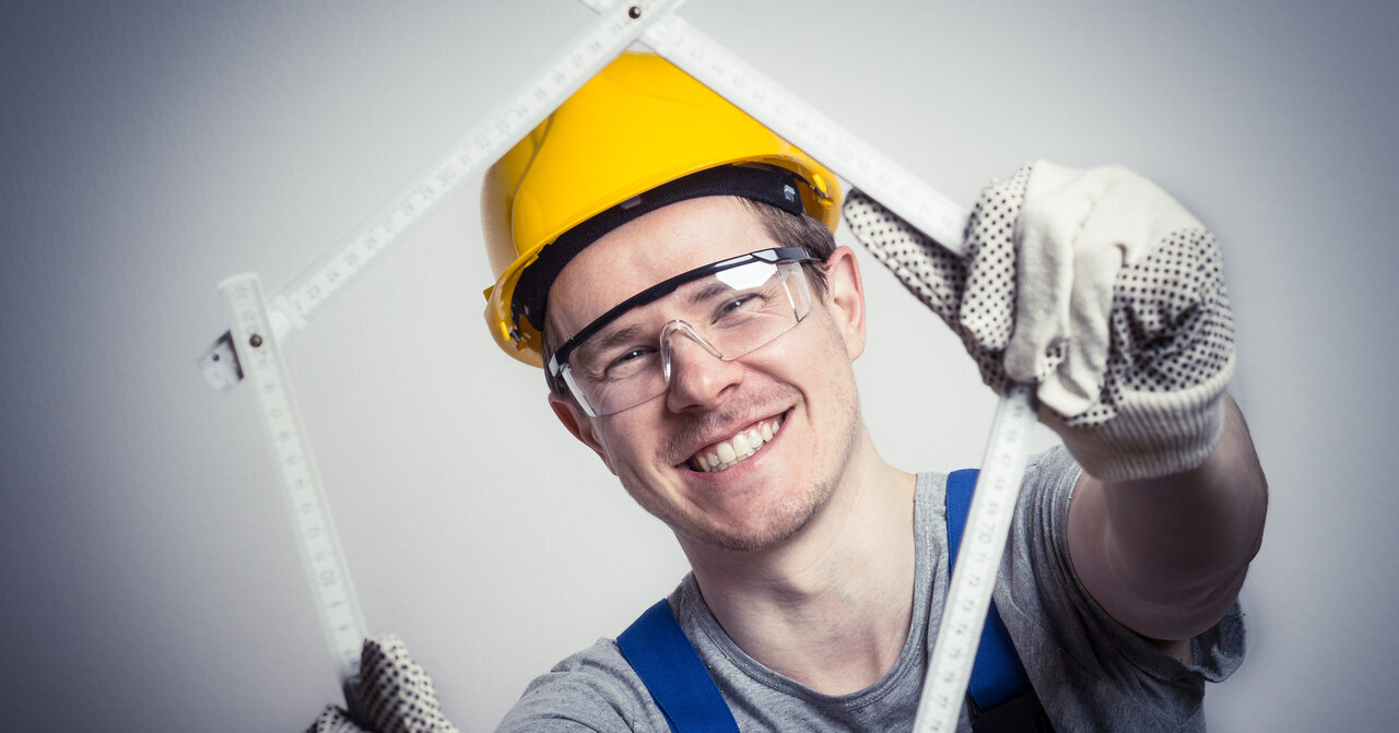Ein Mann mit Schutzbrille und Schutzhelm lächelt durch ein Fünfeck, das er aus einem Zollstock geformt hat.