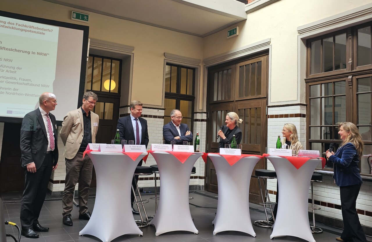 Das Bild zeigt Personen, die an Stehtischen an einem Fachgespräch teilnehmen. Dies sind Eric Thode (Bertelsmann Stiftung), Dr. Peter Brandt (Deutsches Institut für Erwachsenenbildung (DIE)), Staatssekretär Matthias Heidmeier, Roland Schüßler (Regionaldirektion NRW der Bundesagentur für Arbeit), Tanja Nackmayr (unternehmer nrw) und Mareike Richter (DGB NRW) 